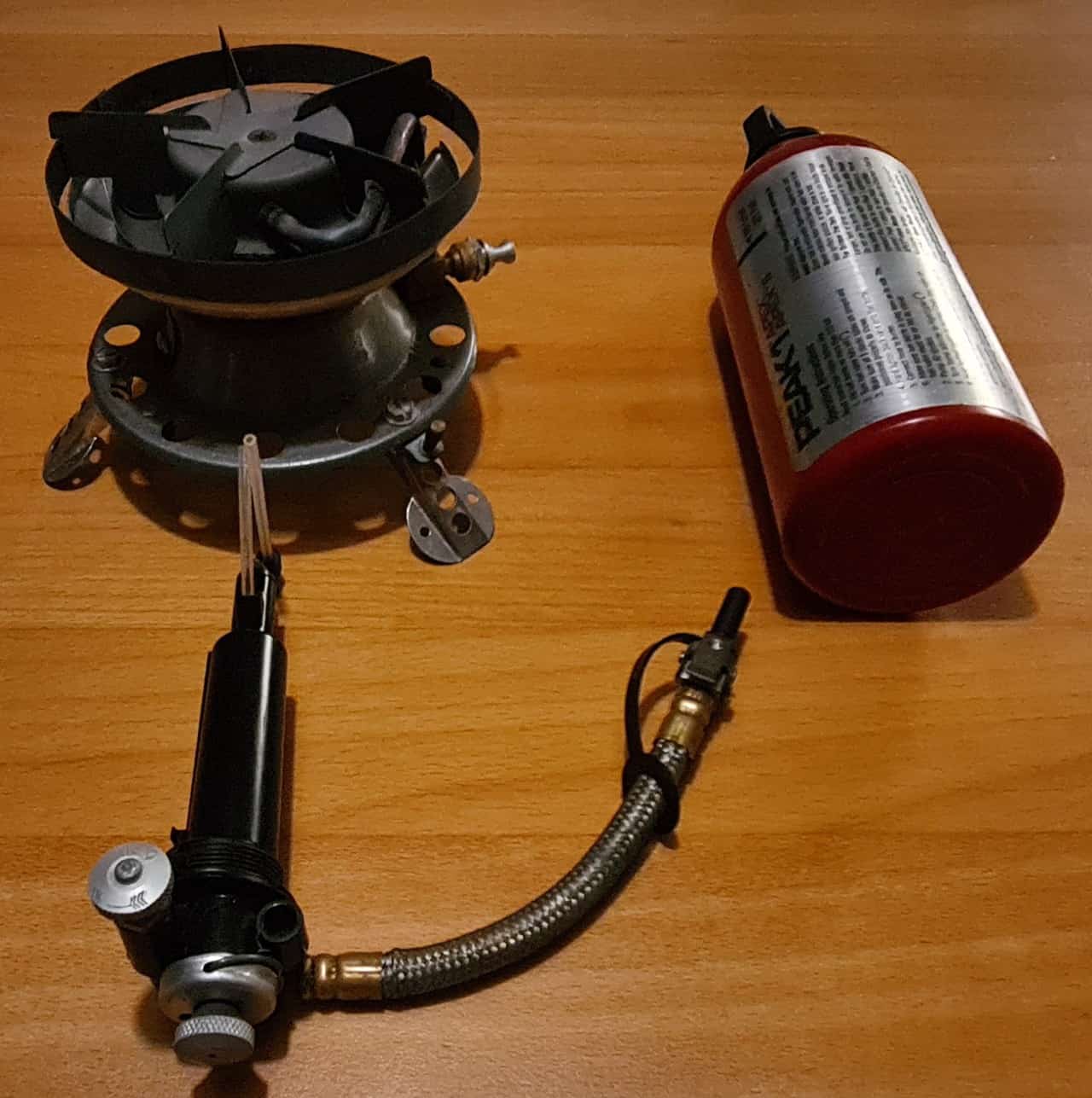 Zerlegter Benzinkocher auf einem Holztisch, links der Brennerkopf mit Windschutz, vorne die Pumpe mit Metallschlauch, rechts eine rote Brennstoffflasche