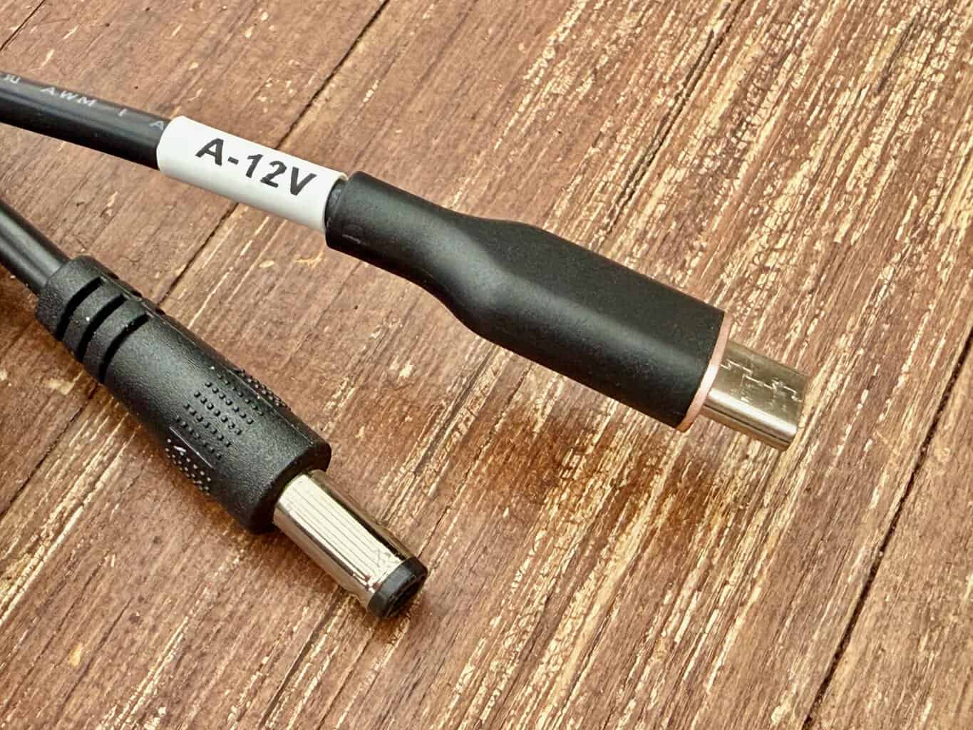 Schwarzes Kabel mit USB-C-Stecker und Hohlstecker auf einem Holztisch, am Kabel ein Etikett A-12V