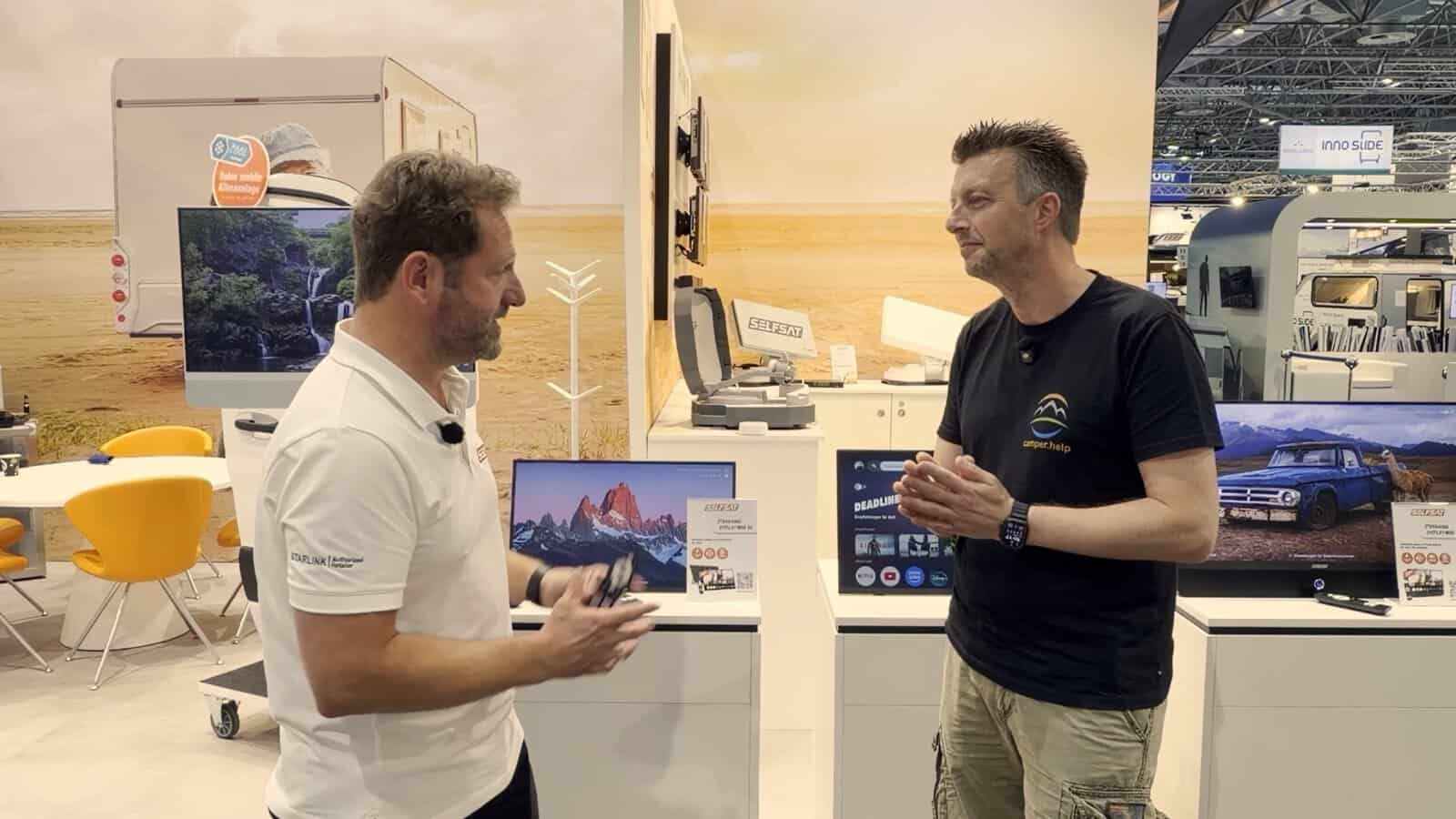 SELFSAT Streaming Displays am Messestand Caravan Salon 2026 Flo von camper.help im Gespräch am SELFSAT-Stand auf dem Caravan Salon 2026, dahinter mehrere Streaming Displays mit Landschaftsmotiven