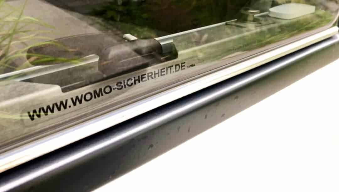 Edelstahlleiste mit der Aufschrift WWW.WOMO-SICHERHEIT.DE unten an der Scheibe eines dunklen Wohnmobilfensters, schräg von der Seite