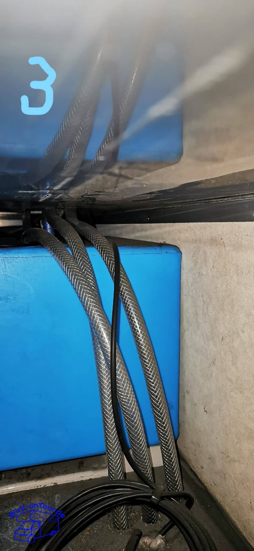 Drei gewebeverstärkte Schläuche und ein schwarzes Kabel laufen in der Heckgarage an einem blauen Kasten vorbei senkrecht nach unten