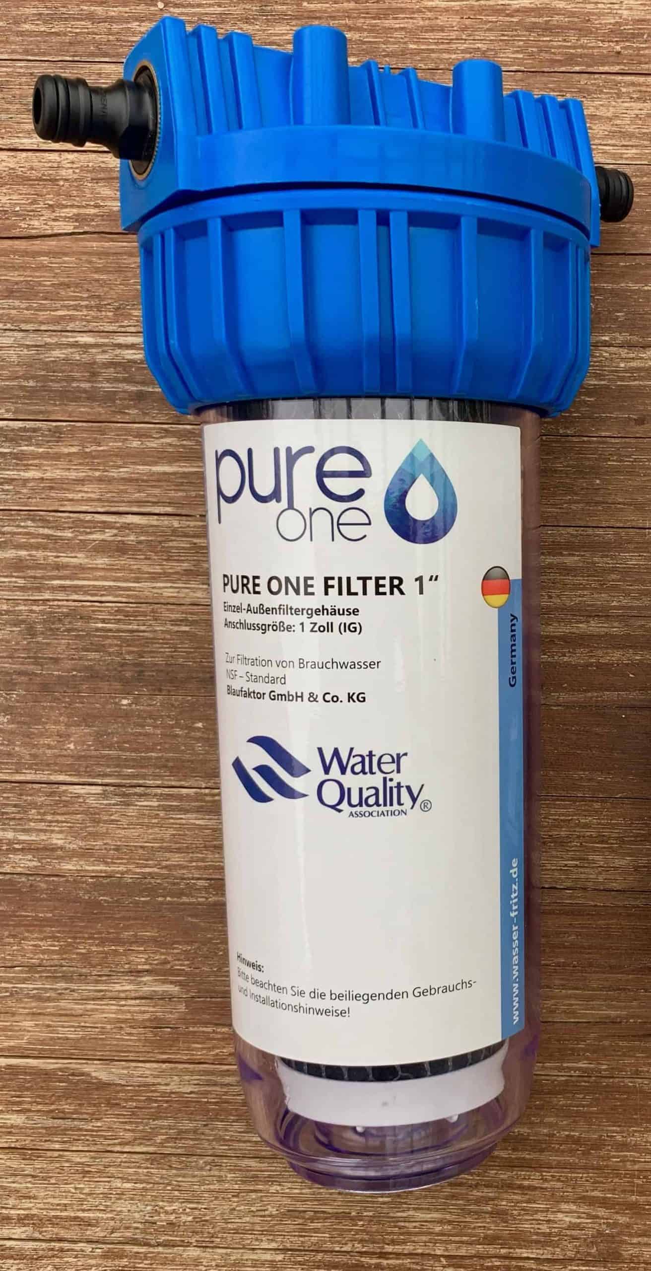Pure One Filtergehäuse mit blauem Deckel und durchsichtiger Glocke, darin eine weiße Filterkartusche mit Pure One Etikett, auf einem Holztisch