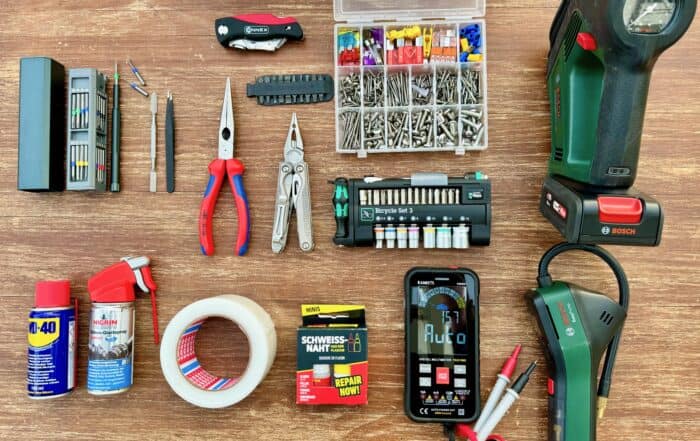 Zehn Werkzeuge auf einem Holztisch ausgelegt: Multimeter, Bosch-Pumpen, Zangen, Bit-Sets, Schraubensortiment, Klebeband und Sekundenkleber