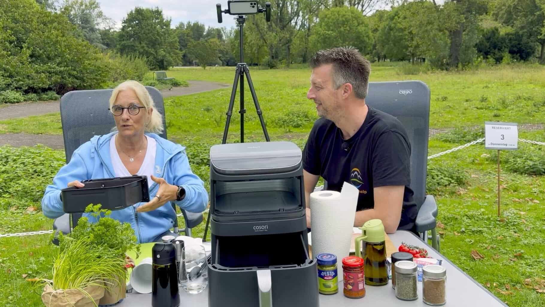 anke-zieht-airfryer-schublade-draussen-heraus Anke hält am Campingtisch auf der Wiese die herausgezogene Schublade des COSORI Airfryers, daneben Flo
