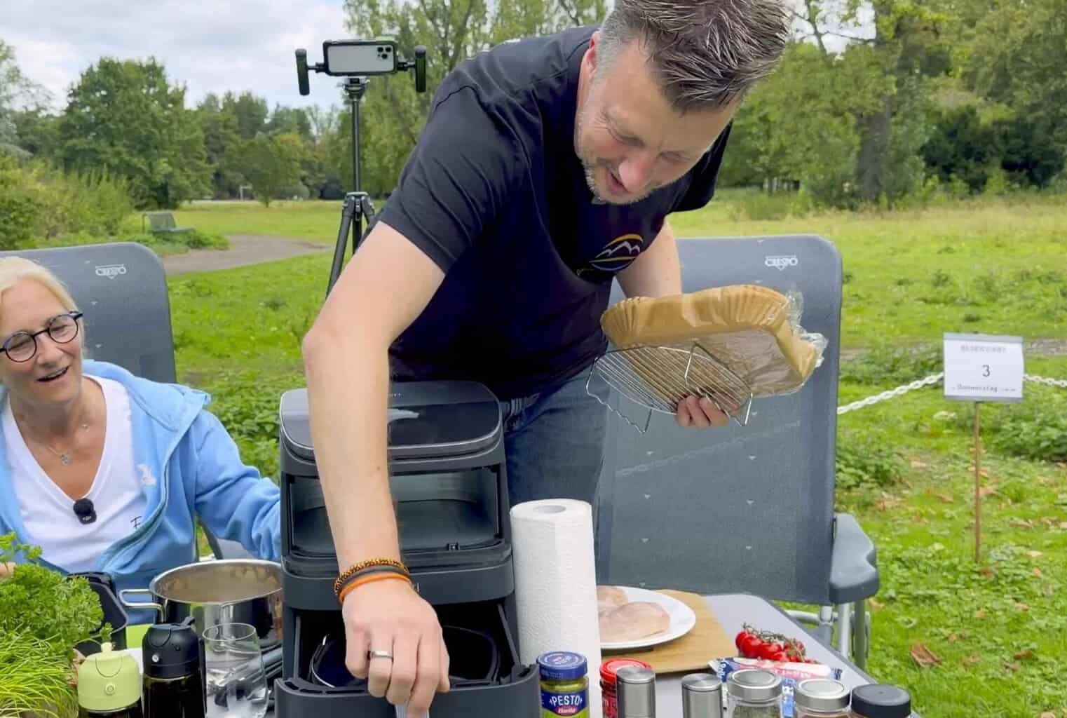 flo-legt-backpapier-auf-gitterrost-in-den-airfryer Flo legt am Campingtisch einen Gitterrost mit zugeschnittenem Backpapier in die Schublade des COSORI Airfryers, links Anke