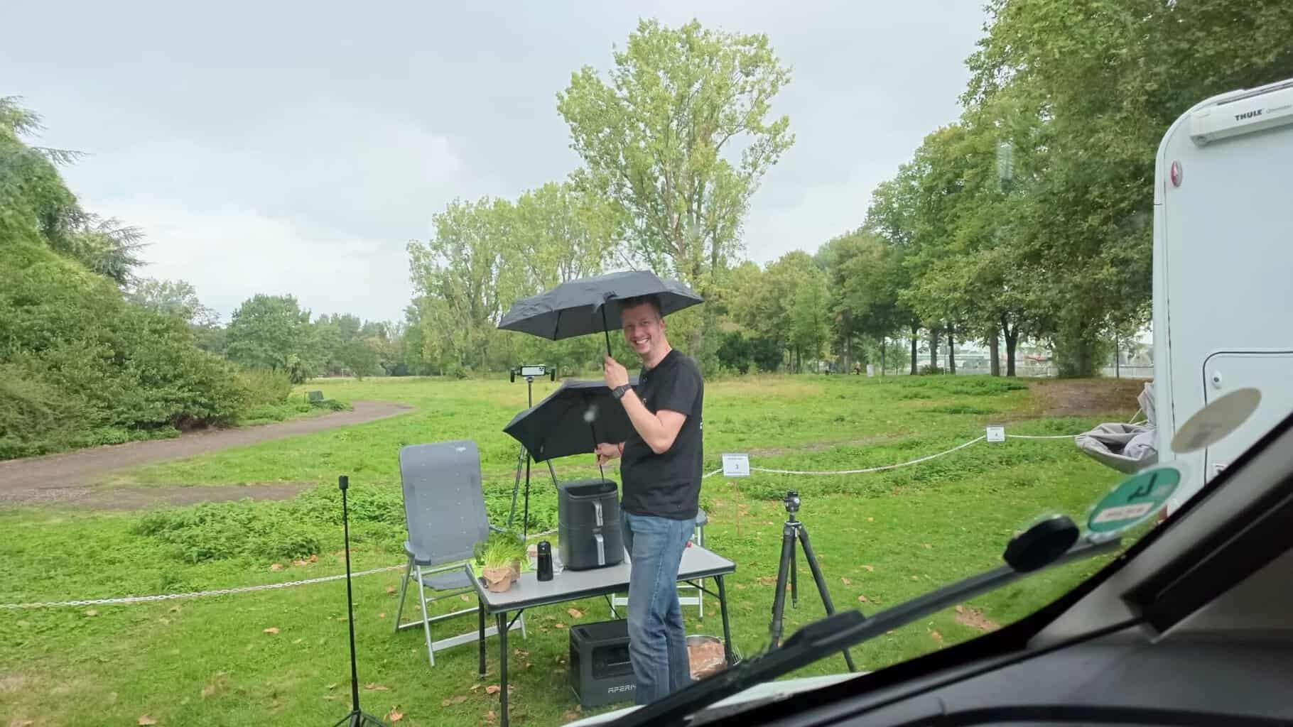 kochen-im-regen-mit-regenschirm Flo steht mit Regenschirm am Campingtisch mit dem Airfryer, im Hintergrund die Wiese